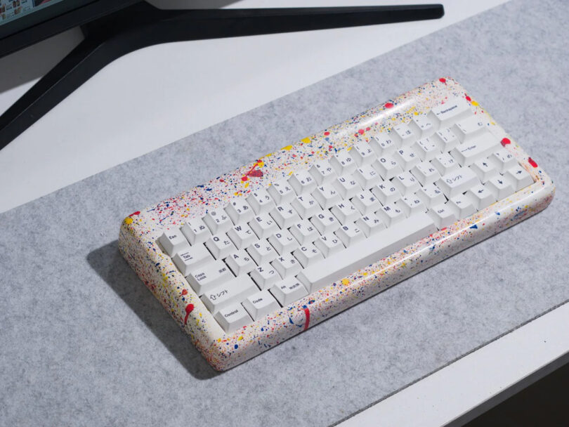 Jawbreaker candy style splattered multicolor paint mechanical keyboard on felt desk pad with monitor legs visible nearby.