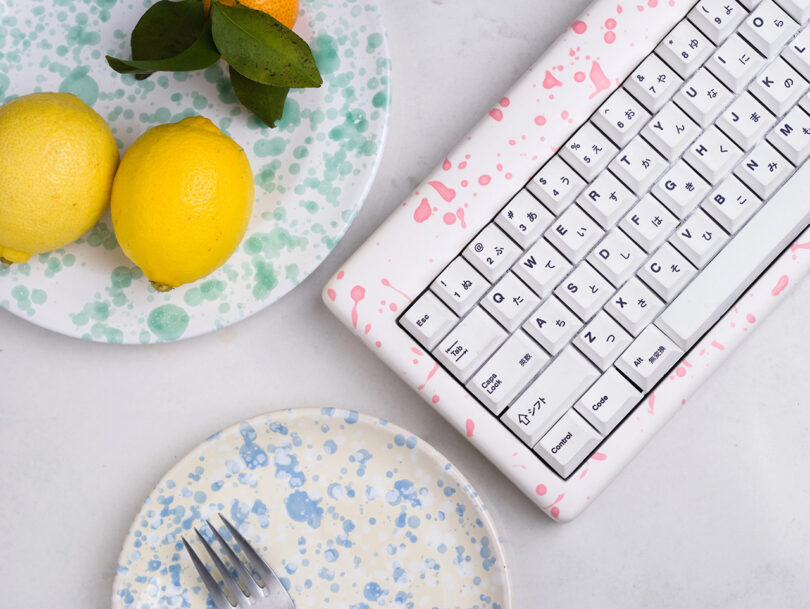 Pink and white resin case paint splattered keyboard near two paint splattered dishes, one with two lemons and a small mandarin citrus on it, the other with a single fork.
