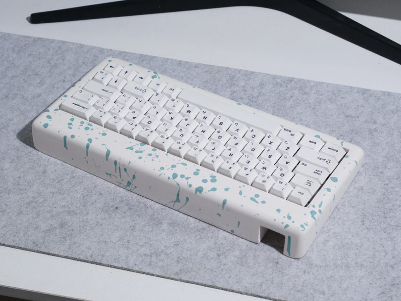 Light blue and white resin case paint splattered keyboard on light gray felt desk pad. Rear side of keyboard case is shown.