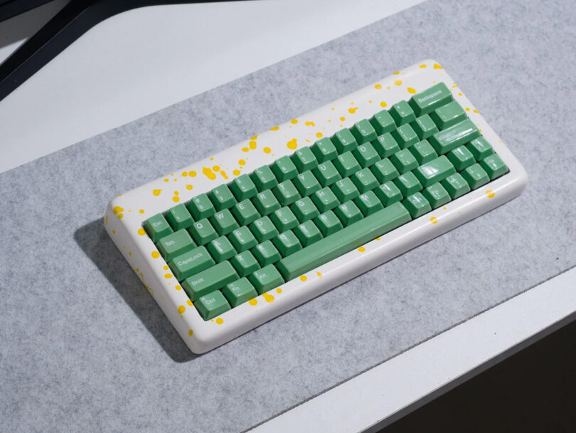 Yellow and white resin case paint splattered keyboard with bright green keycaps, on light gray felt desk pad.