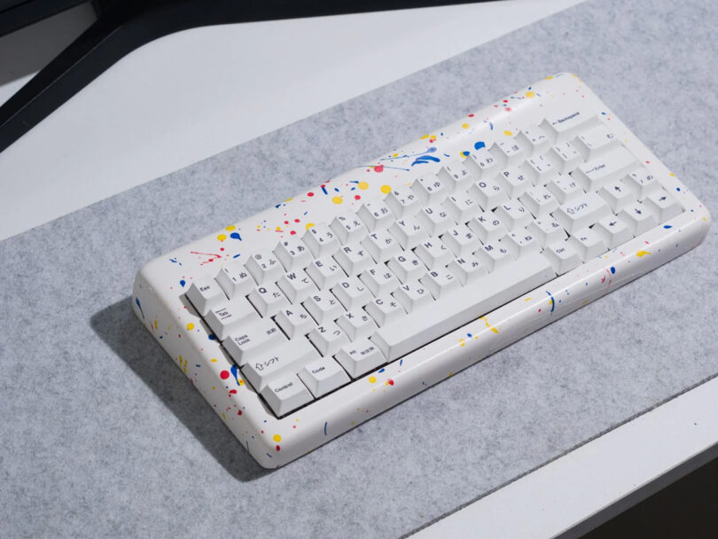 Multicolor and white resin case paint splattered keyboard with white keycaps, on light gray felt desk pad.