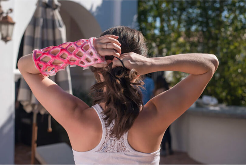 Woman tying back her hair wearing pink and white Cast21 orthopedic sleeve on her arm while outdoors.
