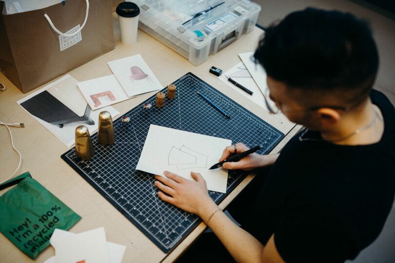 View overhead of someone sketching on top of a studio table cluttered with supplies.