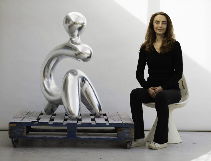 light-skinned woman with long black hair wearing all black sits in a chair next to an abstract silver sculpture