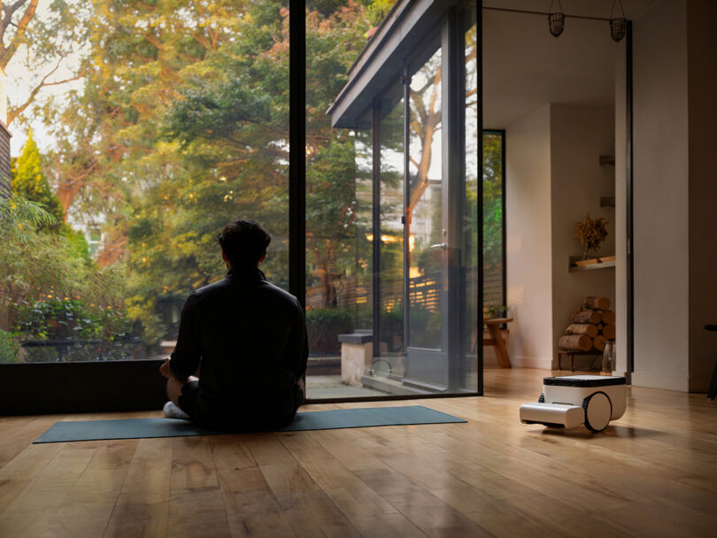 Man on yoga mat meditating staring out a large window while a robot vacuum roves behind him.