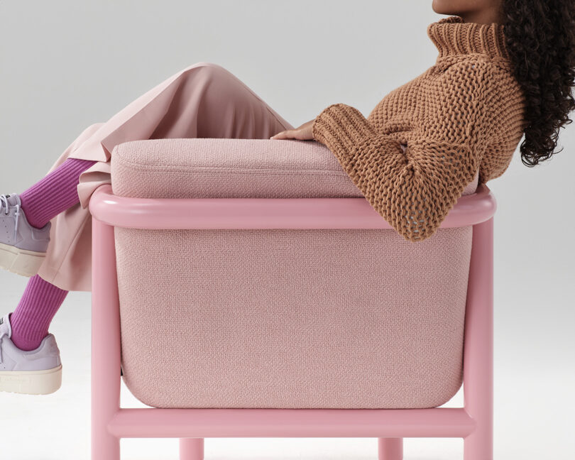 Young woman in cable knit sweater, pink trousers, with pink socks and violet sneakers lounging sideways across all-pink Percy lounge chair.