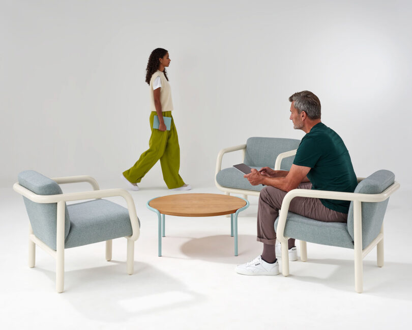 Young woman with blue cover book in hand walking past a man seated in one of three Percy lounge chairs with cream tubular frame and light blue upholstery, with circular table in the center.
