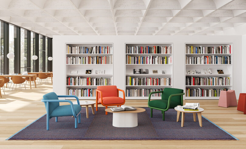 Trio of Percy lounge chair in blue, orange-red, and dark green in the center of a library set across a dark blue rug with bookcases behind it. A row of desks and tables next to floor to ceiling panels of windows are to the left.