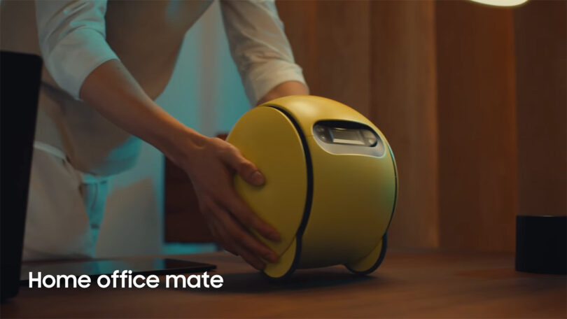 Person placing yellow 3-wheel Samsung AI robot Ballie onto desk to use as a projector