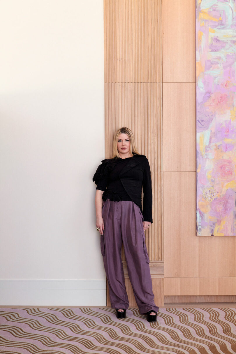 designer Kelly Behun standing in front of wood wall with purple pants and a black blouse