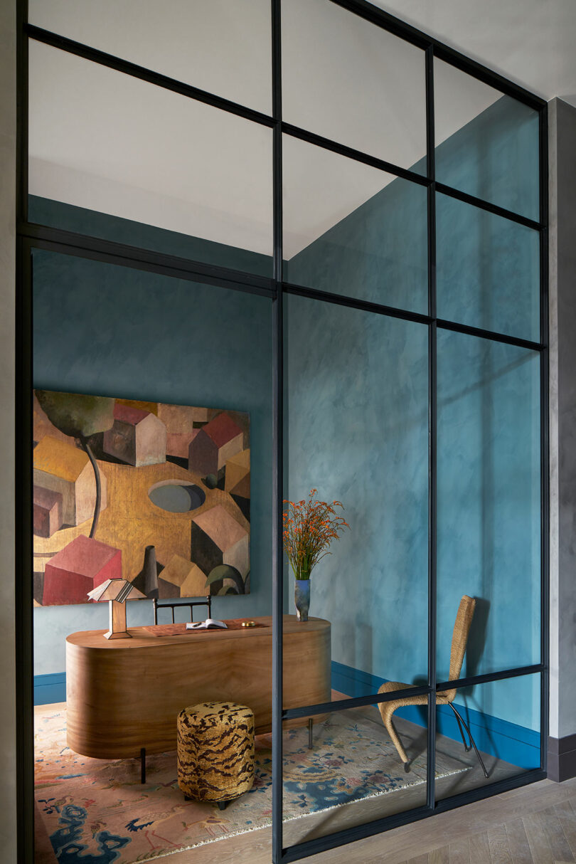 angled view into modern office behind glass wall with modern desk, dark painted blue/green walls and large painting on wall