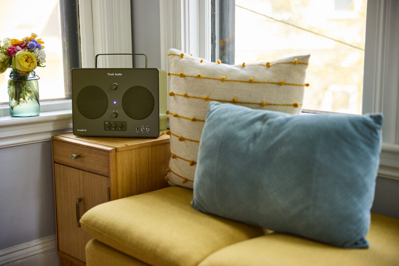 Tivoli Audio SongBook wireless speaker radio in glossy green finish set on a small side end table in a living room near sofa topped with two pillows and jar filled with cut flowers set on a windowsill.