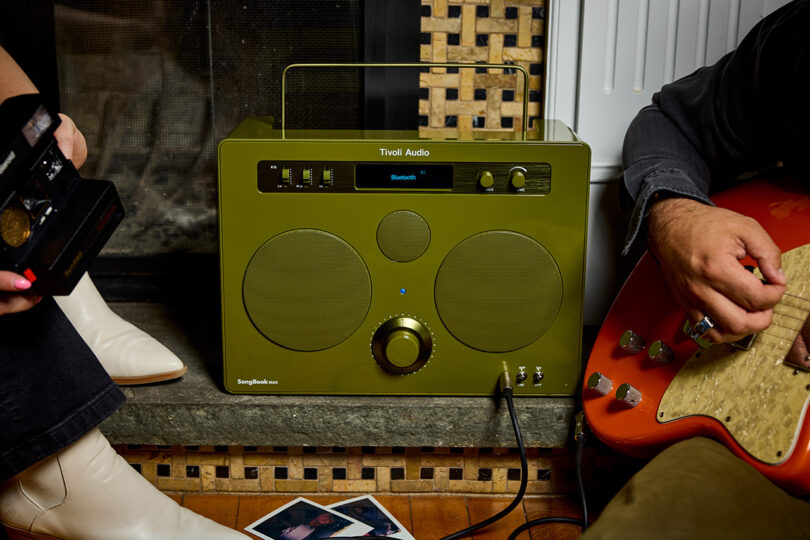 Tivoli Audio SongBook MAX wireless speaker radio in glossy green connected to a guitar a man is strumming to the right, with a woman holding a Polaroid camera to its left.