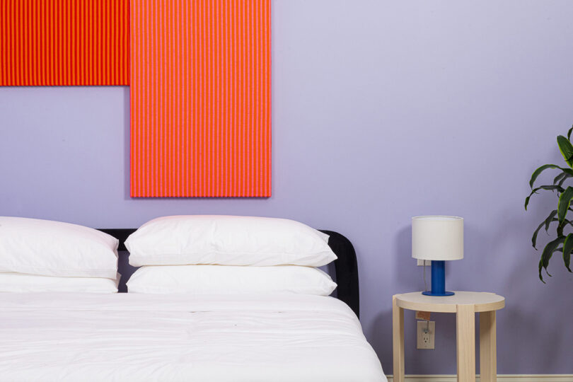 Violet painted wall bedroom inside the YOWIE hotel, with small circular wood side table and blue lamp, and red-orange wall panels placed above the bed.