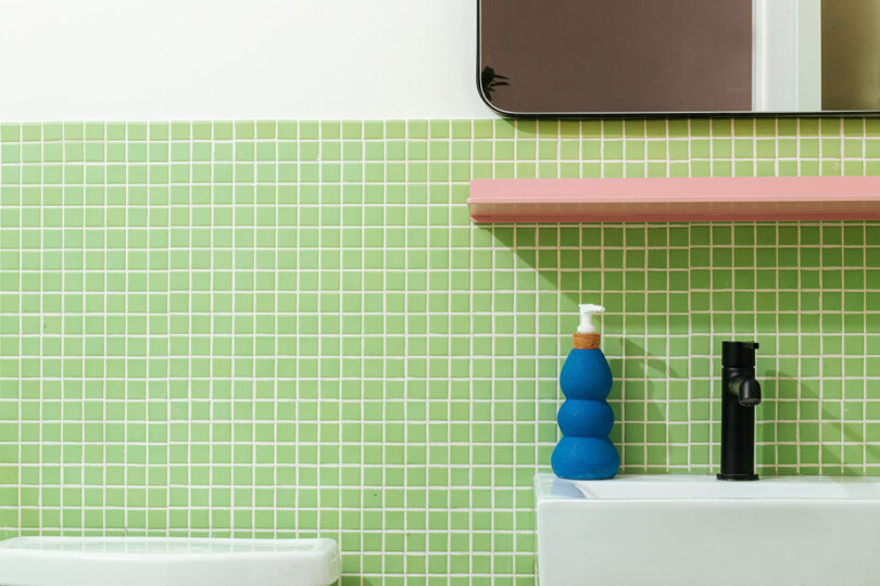 Green mosaic tile bathroom with blue ceramic soap dispenser and pink Shelfology shelf underneath a bathroom mirror.