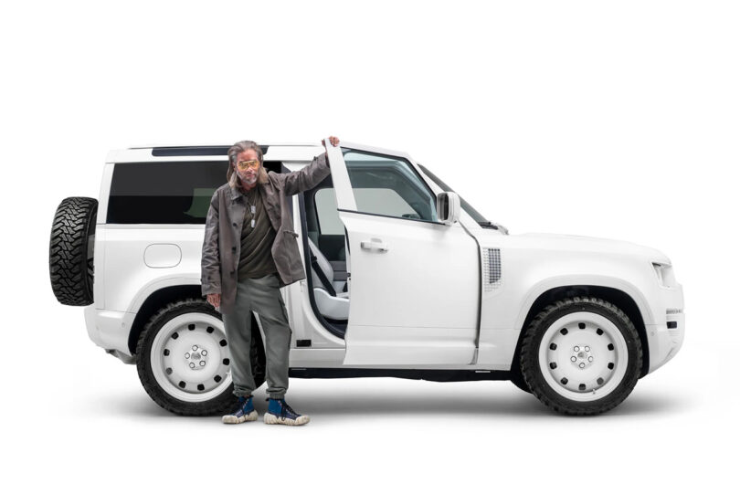 Designer and artist Job Smeets standing next to the passenger side of the Firmship Defender SUV; he's holding the front top corner of the door with his left hand, wearing jeans and a leather jacket.