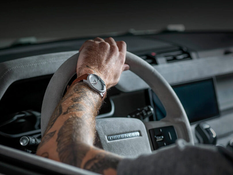 Driver inside Firmship Defender and behind steering wheel wearing a Firmship Aera x P–1 Titanium edition watch