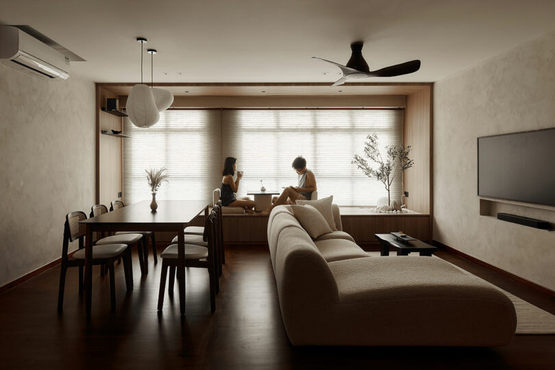 interior view of modern living room and dining room space with sofa and dining table looking towards wall of windows with shades down and two people sitting on the ledge