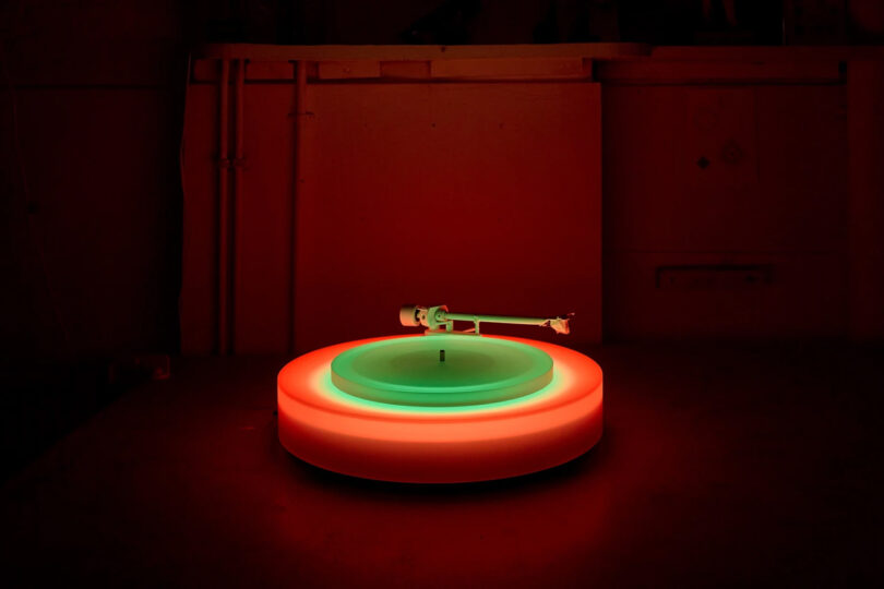 Brian Eno's Turntable II on a table glowing orange, red and green in a dimly red light illuminated room.
