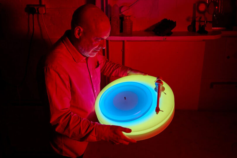 Brian Eno holding up the Turntable II at an angle, the turntables' LEDs glowing red, green and blue across each level. The surrounding room is bathed in red light.