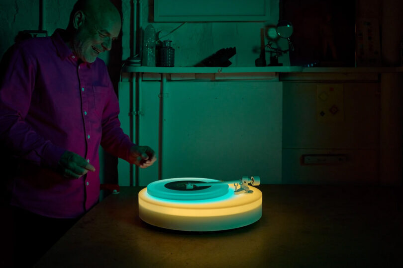 Brian Eno standing over his Turntable II set upon a table glowing green and yellow within a green light illuminated room.