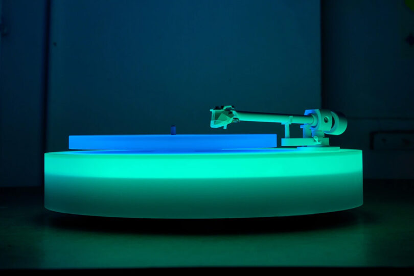 Side view of Brian Eno's Turntable II on a table glowing green and blue showing tone arm and acrylic platter.