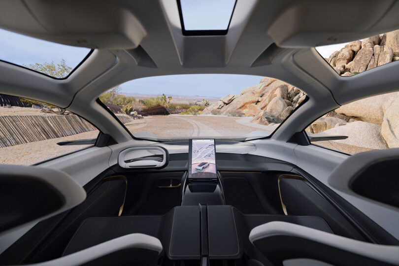 Interior cabin view of the Chrysler Halcyon Concept driving down a desert residence driveway with large boulders visible to the right. Both driver and passenger front seats are unoccupied.