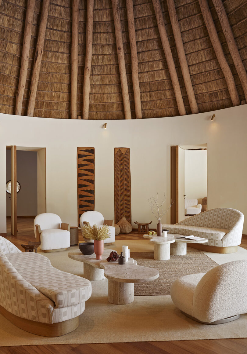 Interior of thatch roof structure furnished with neutral modern furnishings, including sofas, armchairs, and stone coffee and side tables.