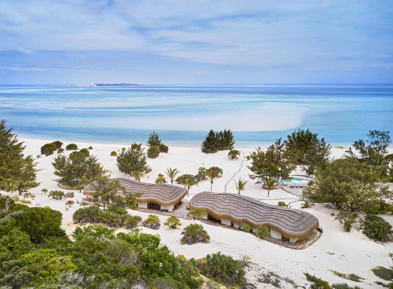 Overhead of the two bungalows Kisawa Residence with an open-air deck, situated on white beach sand and coastal forest facing the azure waters of the Indian Ocean. Another island is visible in the distance across the ocean. 