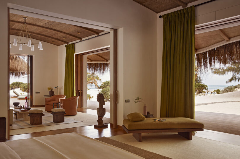 Residência de teto alto e quarto com grandes portas abertas para a praia. Um sofá-cama verde-amarelo claro fica sobre um tapete com duas poltronas e outros móveis visíveis em uma sala adjacente.