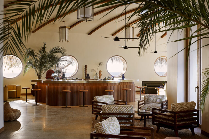Área de bar comum com vigias na parede, 7 bancos, quatro poltronas e grandes palmeiras decorando o interior.