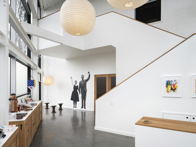 Gift shop section of the Eames Archive with large paper lantern globe lights above and staircase in the background. A print photo of Ray and Charles Eames waving adorns the wall in the background.