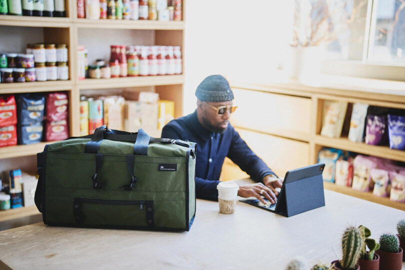 Man with sunglasses and beanie on seated in a small grocery/eatery typing on an iPad with Mission Workshop Mass Transit duffel set across the table to the side of him.