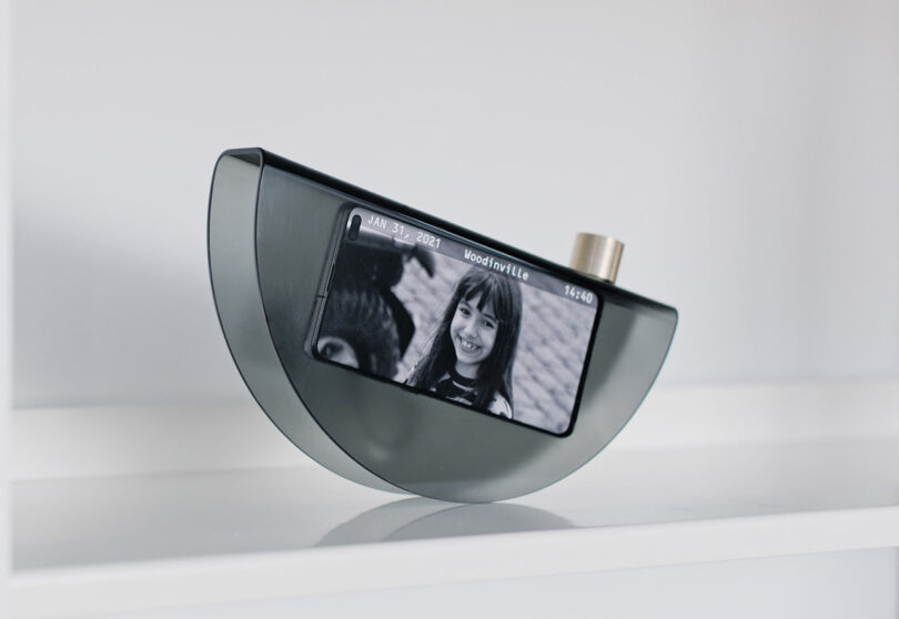 A piece of recycled smoky transparent plastic shaped into an inverted taco shell with a cell phone inside displaying a photo of a young girl smiling standing near a tractor.