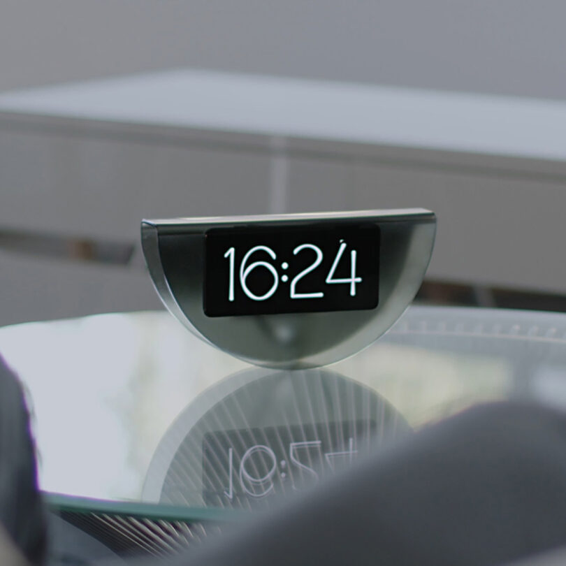 A piece of recycled smoky transparent plastic shaped into an inverted taco shell with a cell phone inside displaying "16:24" in clock mode set across a circular glass top table with metal wire base.