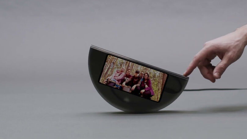 Person's hand pushing on the righthand side of a piece of smoky-transparent plastic shaped into an inverted taco shell with a cell phone inside displaying a photo of a friends seated in an outdoor setting.