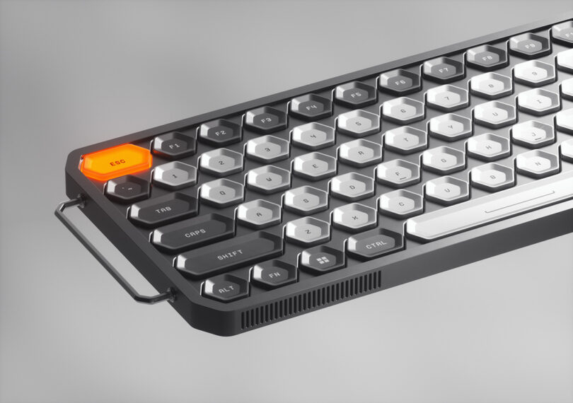 Half left side of the K-Bird mechanical keyboard. ESC key located at upper left of key is backlit orange, with letter keys white and number and function keys in black. Left side is accessorized with small metal handle pull, vents visible along the bottom left side of the keyboard case.
