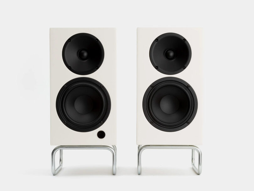 Two white Adsum + ELAC Debut CONNEX DCB41 audio speakers on bent metal leg stands against a white background.