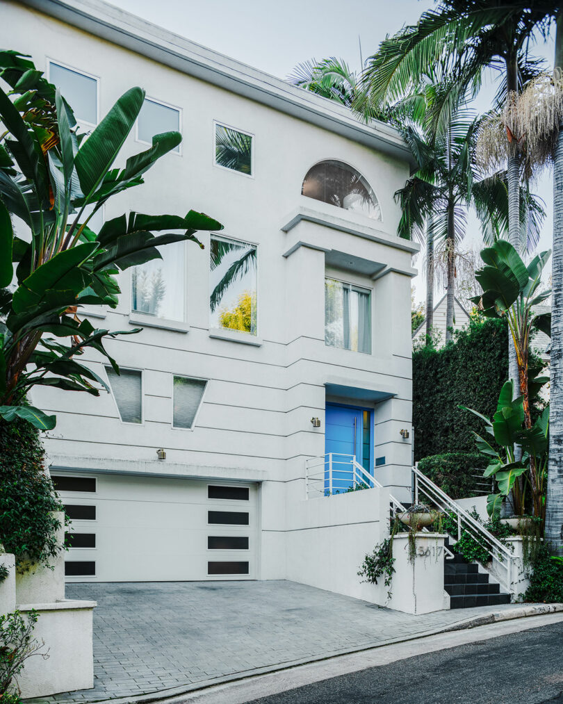 ساختمان مسکونی سفید مدرن با درب ورودی آبی رنگ که با فضای سبز سرسبز احاطه شده است.