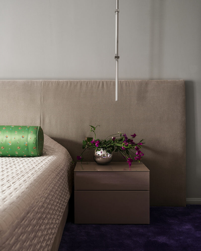 A modern bedroom corner with a beige upholstered headboard, a wooden bedside table, and decorative flowers, complemented by a plush purple carpet.