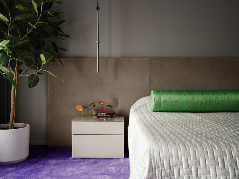 A modern bedroom corner with a beige upholstered headboard, a wooden bedside table, and decorative flowers, complemented by a plush purple carpet.