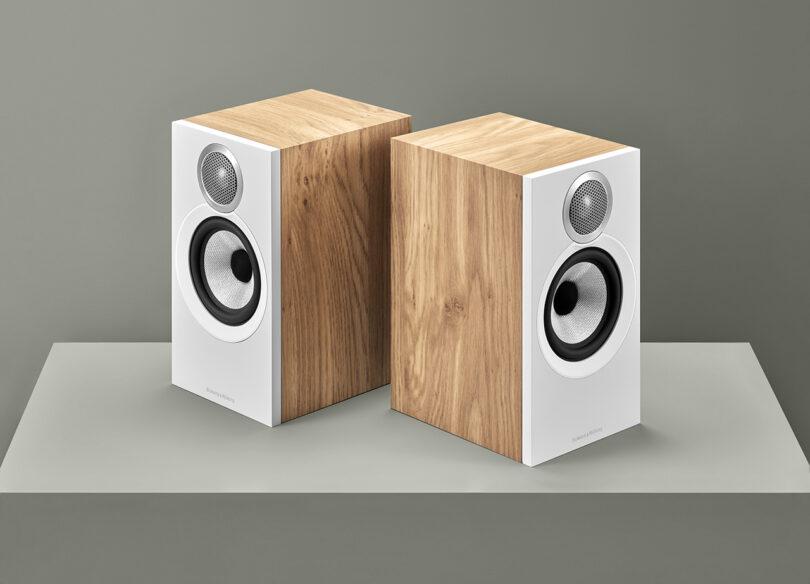 Two Bowers & Wilkins 600 Series bookshelf speakers with a white and oak cabinet finish on a grey-green surface set on a pedestal.