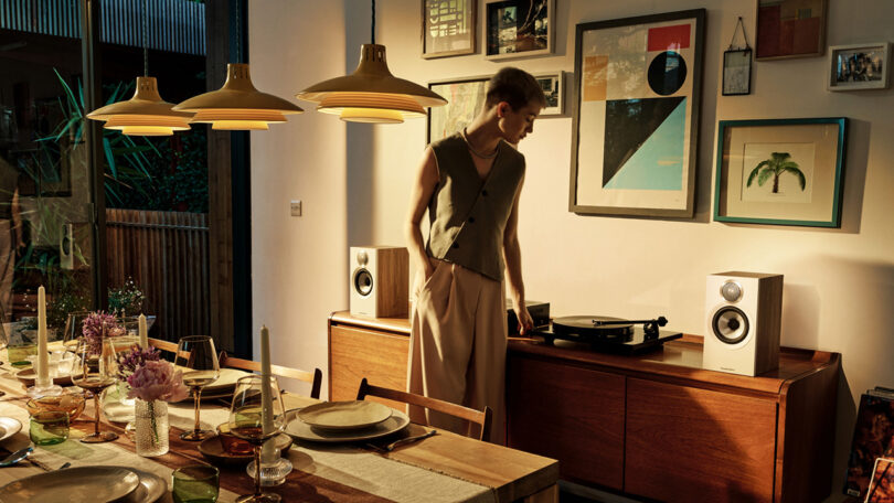 A person standing by a record player and Bowers & Wilkins 600 Series speakers in a warmly lit dining room with a table set for a meal, with gallery wall of framed art in background.