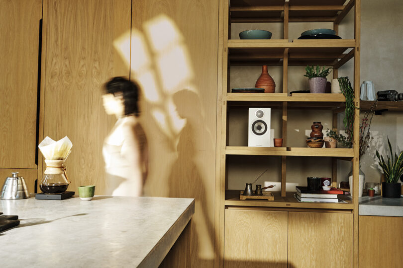 A blurred figure moves through a well-lit, modern kitchen with wooden cabinets and shelves displaying various items, including Bowers & Wilkins 600 Series speakers.