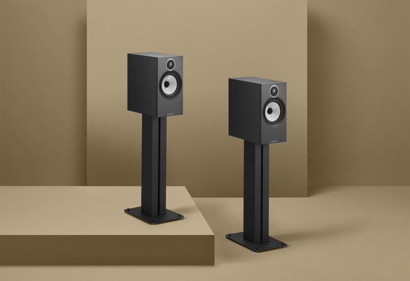 A pair of black Bowers & Wilkins 600 Series speakers on stands against a beige background.