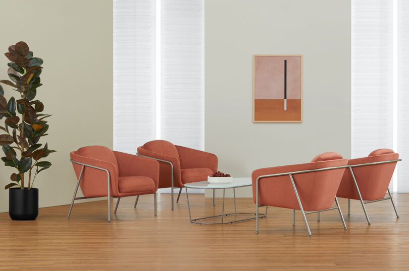 A modern waiting room featuring four coral armchairs, a white coffee table, large windows with sheer curtains, a potted plant, and an abstract painting on the wall.