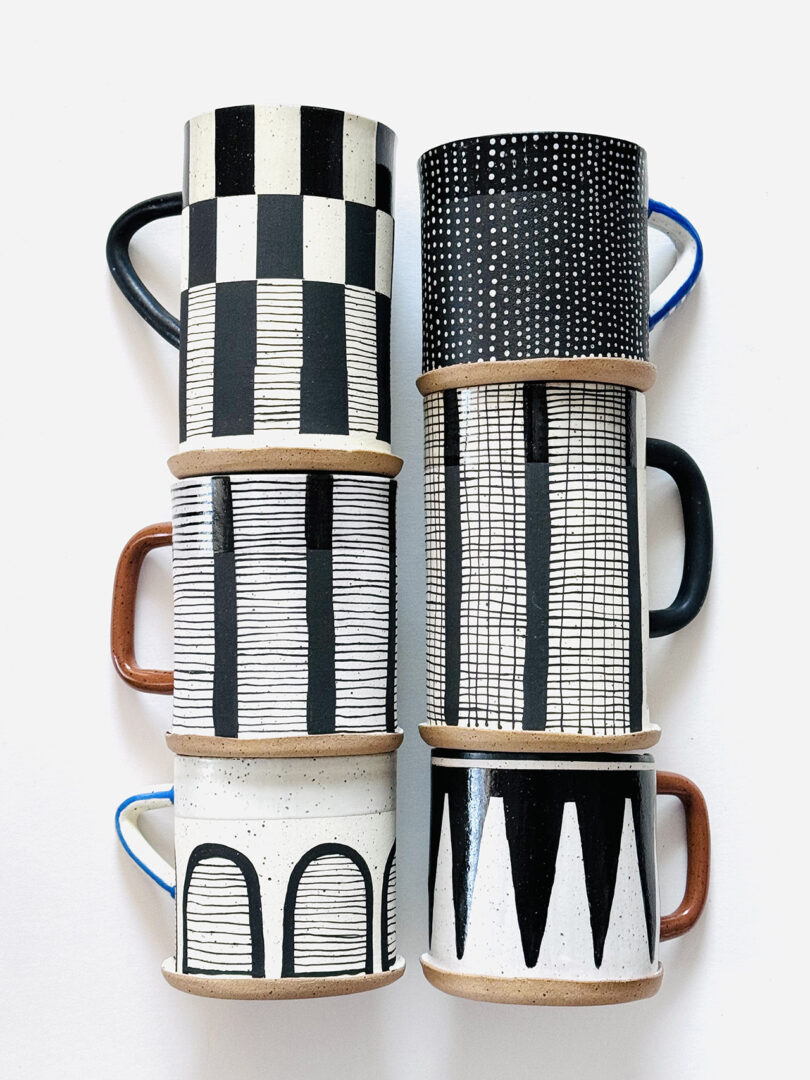 six handmade clay mugs of differing sizes covered in abstract black and white hand-drawn patterns