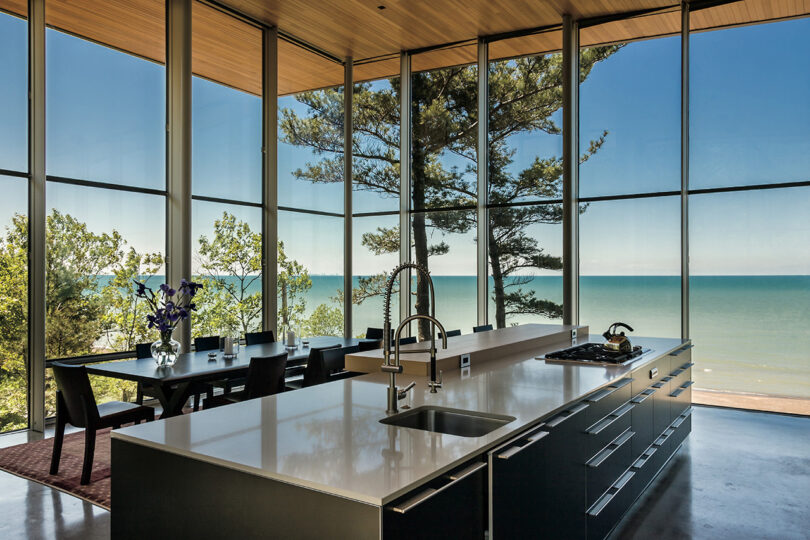 Modern kitchen with floor-to-ceiling windows and a view of the sea and surrounding trees.