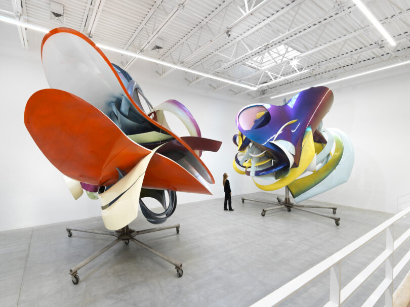 A person observing large, colorful abstract sculptures displayed in a white-walled gallery space.