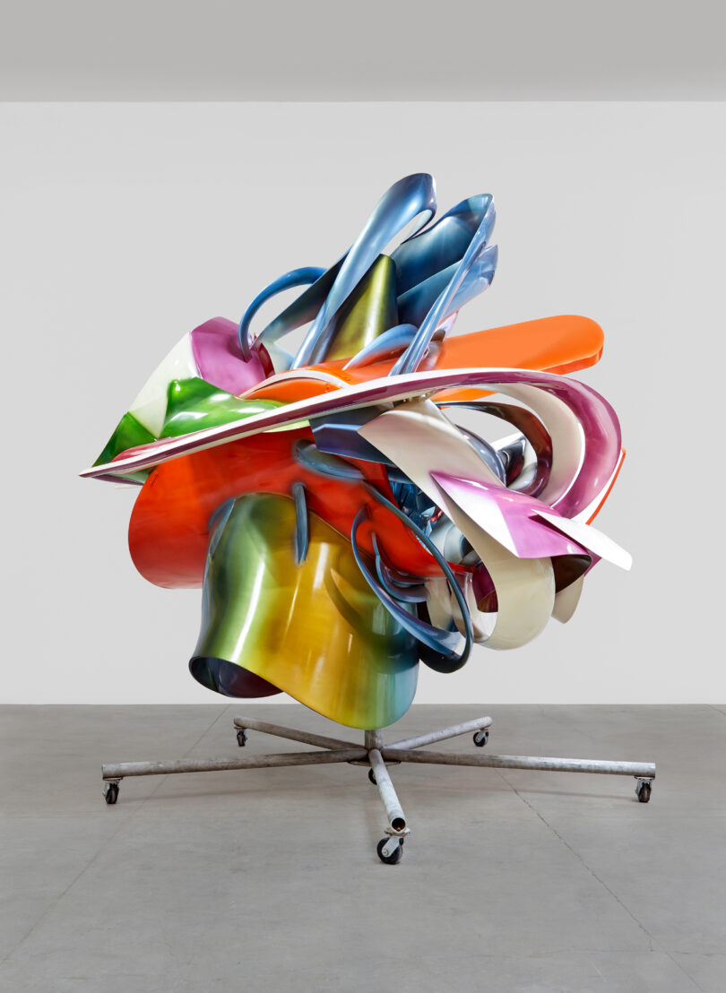 Colorful abstract metal sculpture displayed on a wheeled stand in a gallery setting.